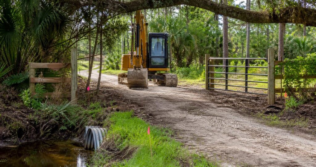 Tracked excavator entering a Florida property through a gate beside a culvert and access markers