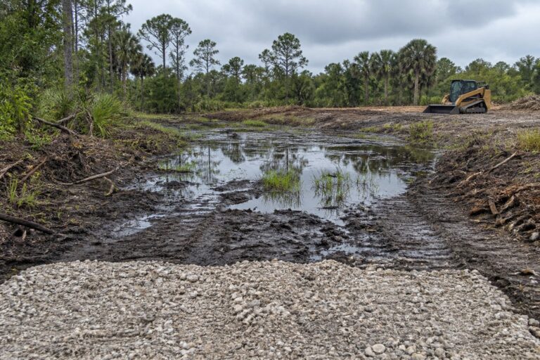 Waterlogged ground beyond a gravel entry with a tracked loader parked in the distance